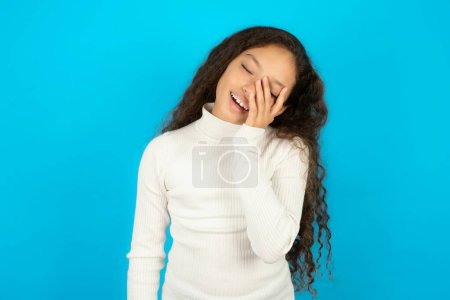 Beautiful kid girl wearing white turtleneck over blue background makes face palm and smiles broadly, giggles positively hears funny joke poses