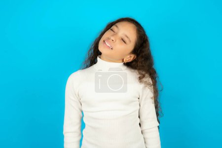 Foto de Positivo Hermosa niña con cuello alto blanco sobre fondo azul con expresión llena de alegría cierra los ojos y se ríe muestra los dientes blancos perfectos - Imagen libre de derechos