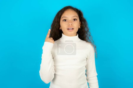Foto de Hermosa niña con cuello alto blanco sobre fondo azul sonriendo y mirando amigable, mostrando el número uno o primero con la mano hacia adelante, cuenta atrás - Imagen libre de derechos