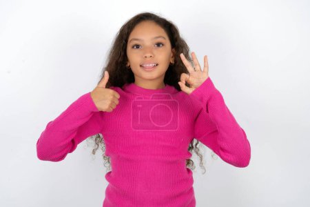 Téléchargez les photos : Belle fille enfant portant col roulé rose sur fond blanc souriant et regardant heureux, insouciant et positif, geste de victoire ou de paix avec une main - en image libre de droit