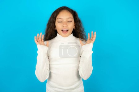 Crazy outraged Beautiful kid girl wearing white turtleneck over blue background screams loudly and gestures angrily yells furiously. Negative human emotions feelings concept