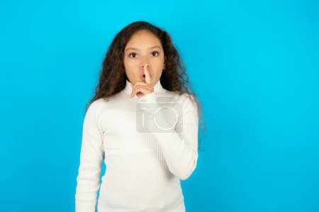 Photo for Surprised Beautiful kid girl wearing white turtleneck over blue background makes silence gesture, keeps finger over lips and looks mysterious at camera - Royalty Free Image