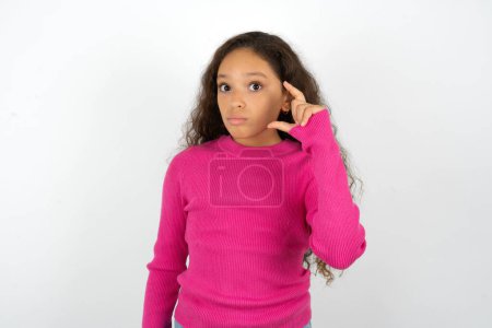 Beautiful kid girl wearing pink turtleneck over white background purses lip and gestures with hand, shows something very little.