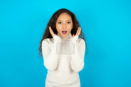 Photo for Surprised happy Beautiful kid girl wearing white turtleneck over blue background glad to see big discounts on clothes, expresses shock, keeps hands near head, jaw dropped. - Royalty Free Image
