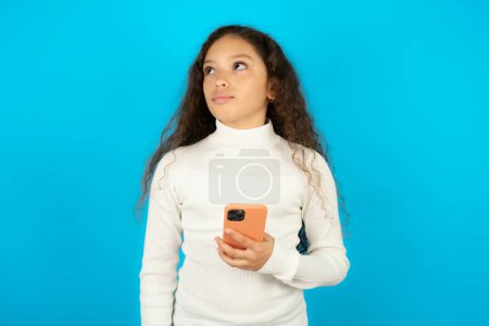 Foto de Hermosa niña con cuello alto blanco sobre fondo azul sostiene teléfono móvil utiliza Internet de alta velocidad y las redes sociales tiene comunicación en línea. Concepto de tecnologías modernas - Imagen libre de derechos