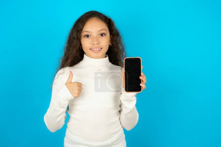 Foto de Hermosa niña con cuello alto blanco sobre fondo azul Mostrar pantalla en blanco smartphone, pulgar hacia arriba recomendar nueva aplicación - Imagen libre de derechos