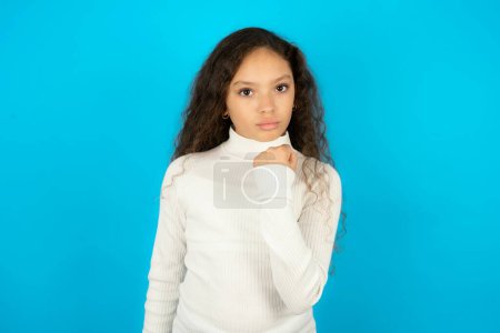 Photo for Beautiful kid girl wearing white turtleneck over blue background shows fist has annoyed face expression going to revenge or threaten someone makes serious look. I will show you who is boss - Royalty Free Image