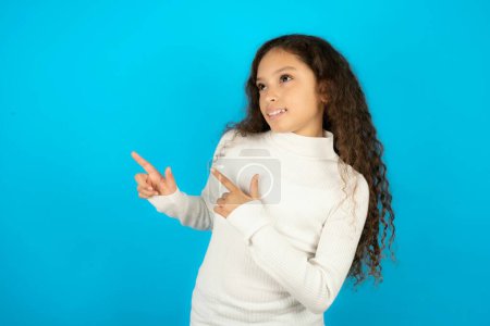 Photo for Optimistic beautiful kid girl wearing white turtleneck over blue background points with both hands and  looking at empty space. - Royalty Free Image