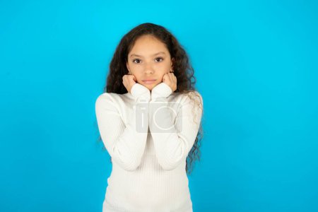 Photo for Portrait of sad beautiful kid girl wearing white turtleneck over blue background hands face - Royalty Free Image