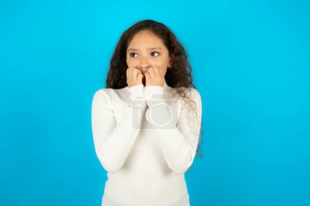 Photo for Terrified Beautiful kid girl wearing white turtleneck over blue background looks empty space home alone moonless night - Royalty Free Image