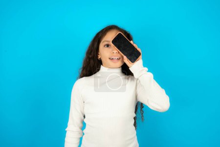 Foto de Hermosa niña con cuello alto blanco sobre fondo azul sosteniendo teléfono inteligente moderno que cubre un ojo mientras sonríe - Imagen libre de derechos