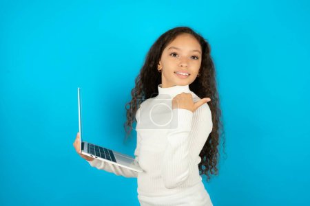Photo for Beautiful kid girl wearing white turtleneck over blue background directing empty space  hold laptop - Royalty Free Image