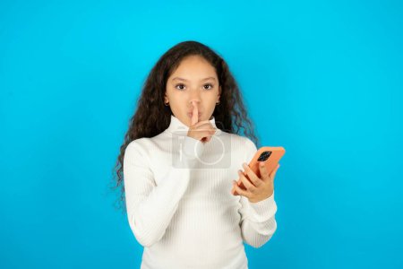 beautiful kid girl wearing white turtleneck over blue background holding modern gadget ask not tell secrets