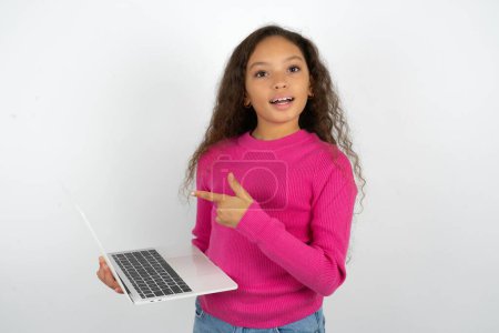 Foto de Shocked Hermosa niña con cuello alto rosa sobre fondo blanco señalando el dedo dispositivo moderno - Imagen libre de derechos