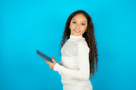 Foto de Foto de optimista Hermosa niña con cuello alto blanco sobre fondo azul mantenga tableta - Imagen libre de derechos