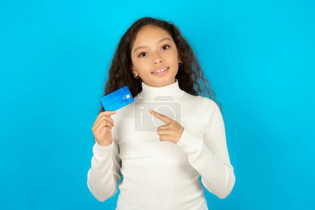 Téléchargez les photos : Portrait photo de belle fille enfant portant col roulé blanc sur fond bleu faire l'achat avec pointant du doigt carte bancaire de crédit - en image libre de droit