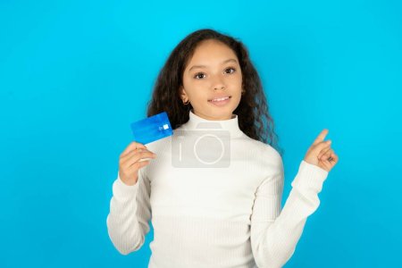 Foto de Sonriente Hermosa niña con cuello alto blanco sobre fondo azul mostrando tarjeta de débito señalando el dedo espacio vacío - Imagen libre de derechos