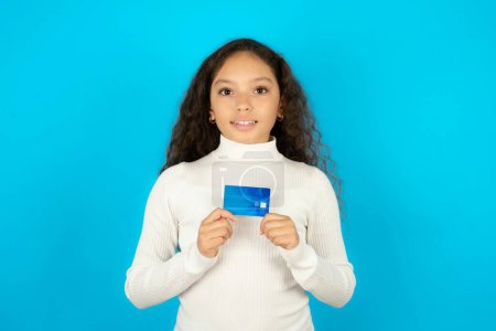 Foto de Foto de niña hermosa alegre usando cuello alto blanco sobre fondo azul mantenga tarjeta de débito mirada espacio vacío - Imagen libre de derechos