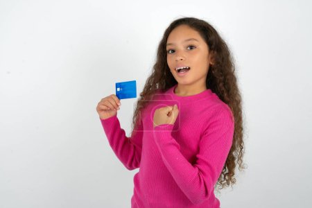 Photo for Excited happy positive cheerful smiling Beautiful kid girl wearing pink turtleneck over white background hold credit card raise fist in victory - Royalty Free Image