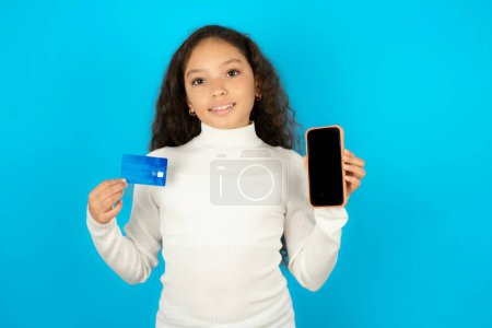Photo for Beautiful kid girl wearing white turtleneck over blue background opened bank account, holding smartphone and credit card, smiling, recommend use online shopping application - Royalty Free Image