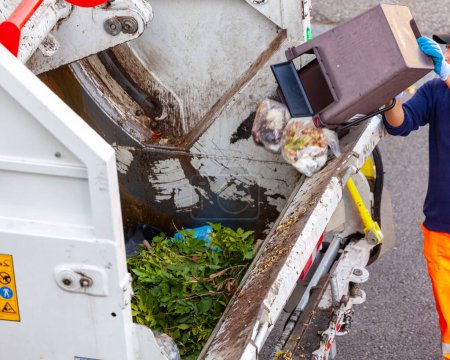Ecological worker empties bucket of organic waste into garbage truck.