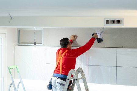 Téléchargez les photos : Travailleur de placoplâtre installe un mur de placoplâtre sur les armoires de cuisine pour couvrir le tuyau d'aspiration de la hotte. - en image libre de droit