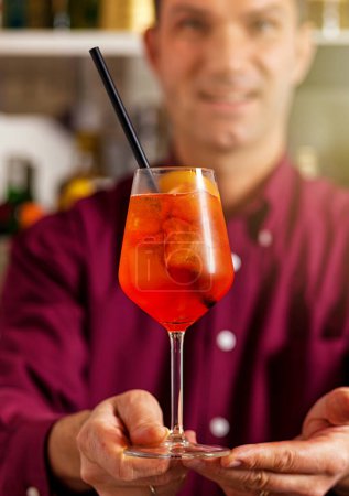 Téléchargez les photos : Barman souriant dans une chemise violette vibrante servant un spritz apéro rafraîchissant dans un bar - en image libre de droit