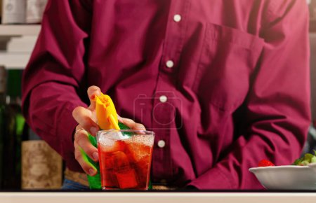 Großaufnahme der Hände eines Mannes, der an einer Bar mit einem Limonadensiphon einen leuchtend roten Cocktail mixt.