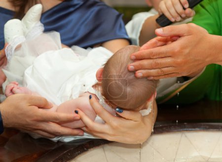 Prêtre verse de l'eau sur la tête d'une petite fille pendant que ses parents la tiennent sur une fontaine baptismale