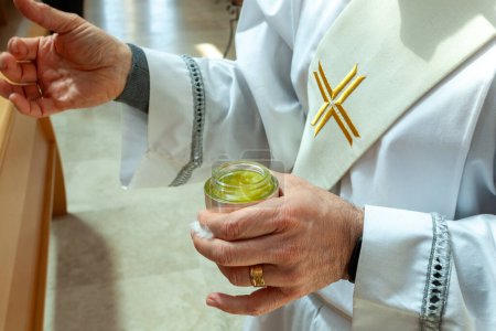 Prêtre portant un vêtement religieux blanc tenant un bocal d'huile de baptême lors d'une cérémonie religieuse