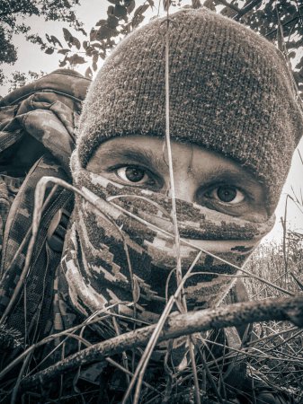 Téléchargez les photos : Man whose face is obscured by balaclava lies prone on the earth deep in hostile territory. Intensity and dedication of special forces operative as he conducts reconnaissance operations on the ground. - en image libre de droit