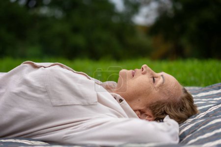 Photo for Elderly woman lying on a plaid in the park with her eyes closed resting and relaxing with nature - Royalty Free Image