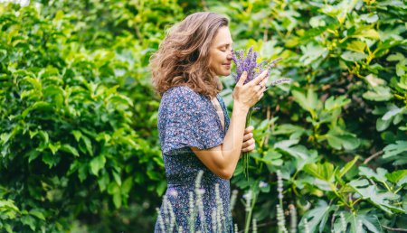 Foto de Joven mujer feliz en un vestido de pie en el jardín con un ramo de lavanda e inhalando el aroma de las flores - Imagen libre de derechos