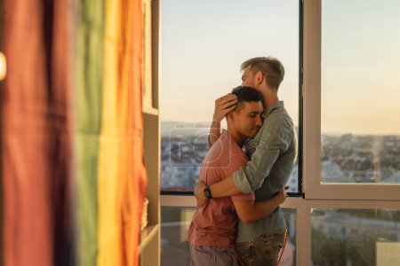 Foto de Joven linda pareja gay abrazándose en un apartamento cerca de la ventana con vistas a la ciudad. Familia LGBT - Imagen libre de derechos
