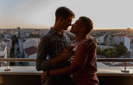 Foto de Joven feliz linda pareja gay abrazándose en balcón con vistas a la ciudad y puesta de sol - Imagen libre de derechos