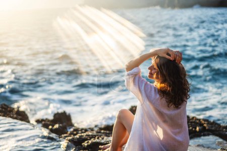 Téléchargez les photos : Jeune femme joyeuse et détendue dans une chemise blanche profitant d'un merveilleux moment de vie tout en étant assis sur le bord de la mer - en image libre de droit