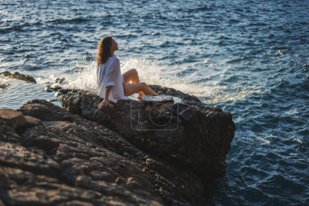 Téléchargez les photos : Jeune femme joyeuse et détendue dans une chemise blanche profitant d'un merveilleux moment de vie tout en étant assis sur le bord de la mer - en image libre de droit