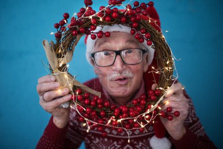 Portrait of happy senior man with Santa Claus hats in studio shoot, looking trough Christmas wreath.