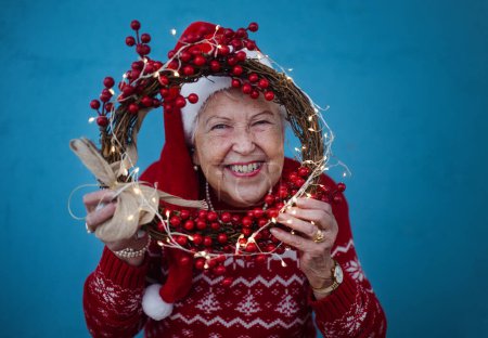 Retrato de mujer mayor feliz con sombreros de Santa Claus en sesión de estudio, mirando a través de la corona de Navidad.