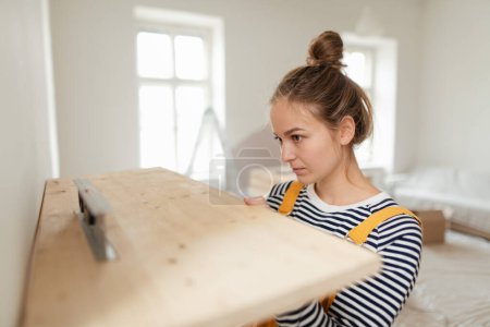 Photo for Young woman remaking her apartment, measuring wall with spirit level and hanging a shelf. - Royalty Free Image