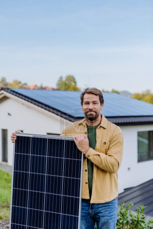Foto de Hombre maduro sosteniendo un panel solar cerca de su casa con paneles solares en el techo. Energía alternativa, ahorro de recursos y concepto de estilo de vida sostenible. - Imagen libre de derechos