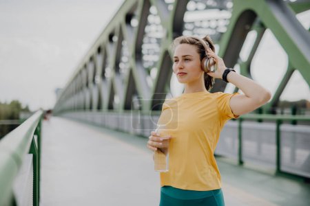 Young woman drinking water and listening the music trough headphones, during jogging in city, healthy lifestyle and sport concept.