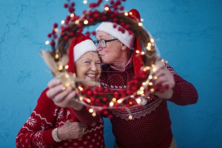 Foto de Retrato de feliz pareja de ancianos con sombreros de Santa Claus en sesión de estudio, mirando a través de la corona de Navidad. - Imagen libre de derechos