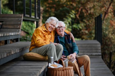 Foto de Feliz pareja de ancianos descansando cerca del lago después de caminar y mirando un teléfono inteligente. - Imagen libre de derechos