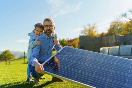 Photo for Father with his little daughter catching sun at solar panel,charging at the backyard. Alternative energy, saving resources and sustainable lifestyle concept. - Royalty Free Image