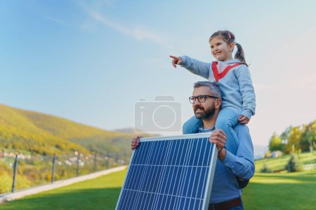 Father with his little daughter on piggyback, catching sun at solar panel,charging it at the backyard. Alternative energy, saving resources and sustainable lifestyle concept.