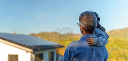 Photo for Rear view of dad holding her little girl in arms and looking at their house with solar panels.Alternative energy, saving resources and sustainable lifestyle concept. - Royalty Free Image