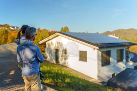 Rear view of dad holding her little girl in arms and looking at their house with solar panels.Alternative energy, saving resources and sustainable lifestyle concept.