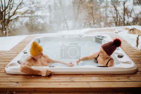 Photo for Senior couple in kintted cap enjoying together outdoor bathtub and clinking glasses at their terrace during a cold winter day. - Royalty Free Image