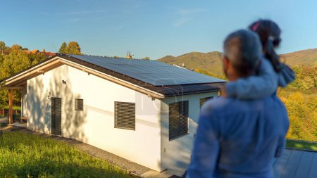 Rear view of dad holding her little girl in arms and looking at their house with solar panels.Alternative energy, saving resources and sustainable lifestyle concept.